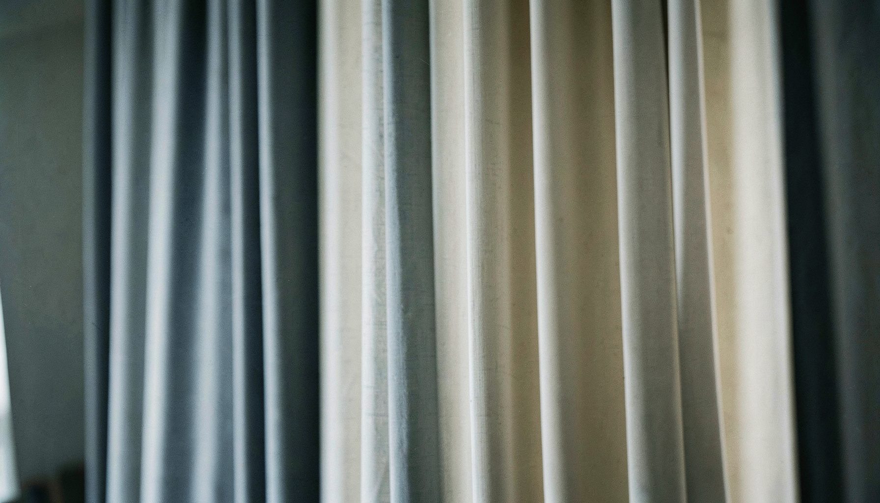 Wide soft-focus view of pleated fabric drapery panels hanging in muted blue-gray and cream tones, gentle folds catching diffused light, calm and understated interior mood